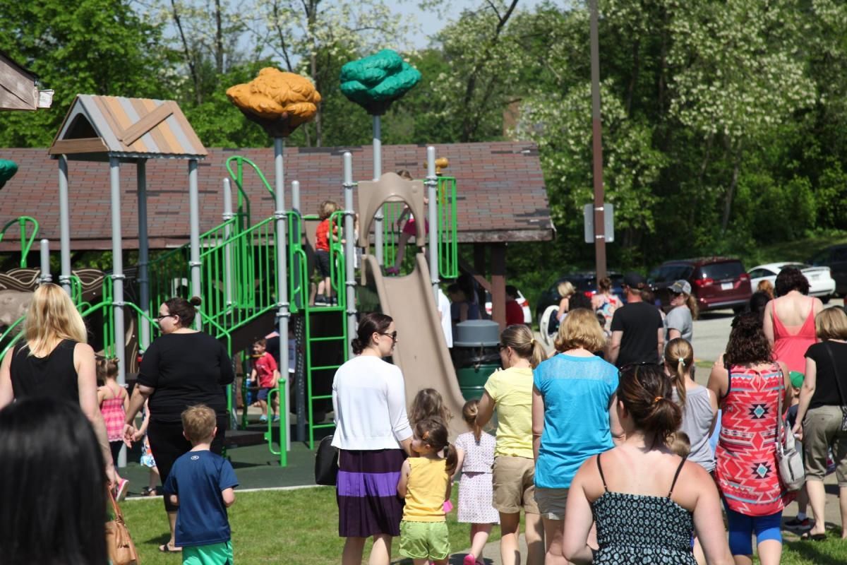 Crowded playground with children and adults near a green slide and play structure on a sunny day