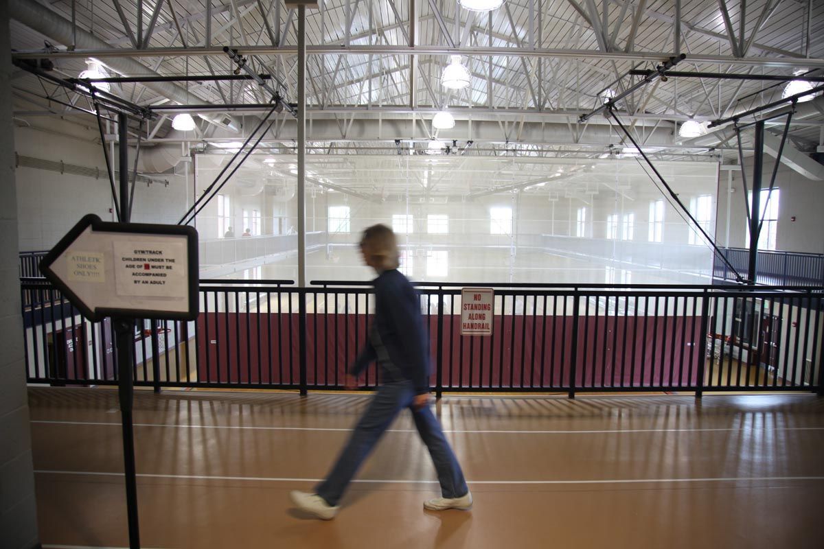 Person walking across an indoor gym with a wooden floor and basketball court in the background.