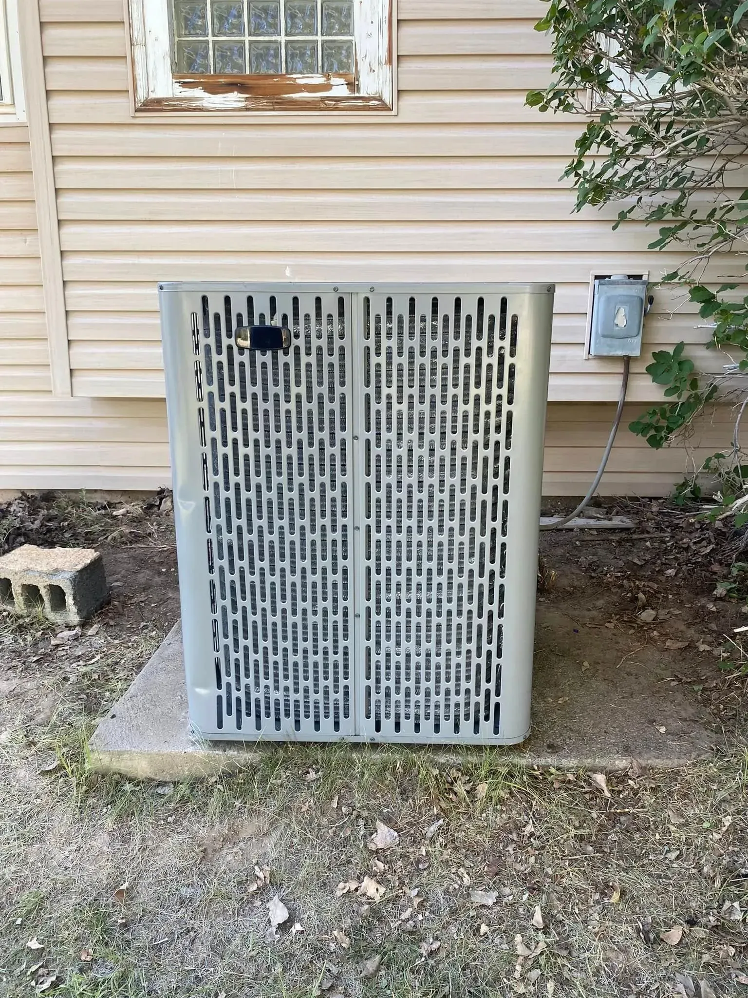 An air conditioning unit outside a house with a weathered window.