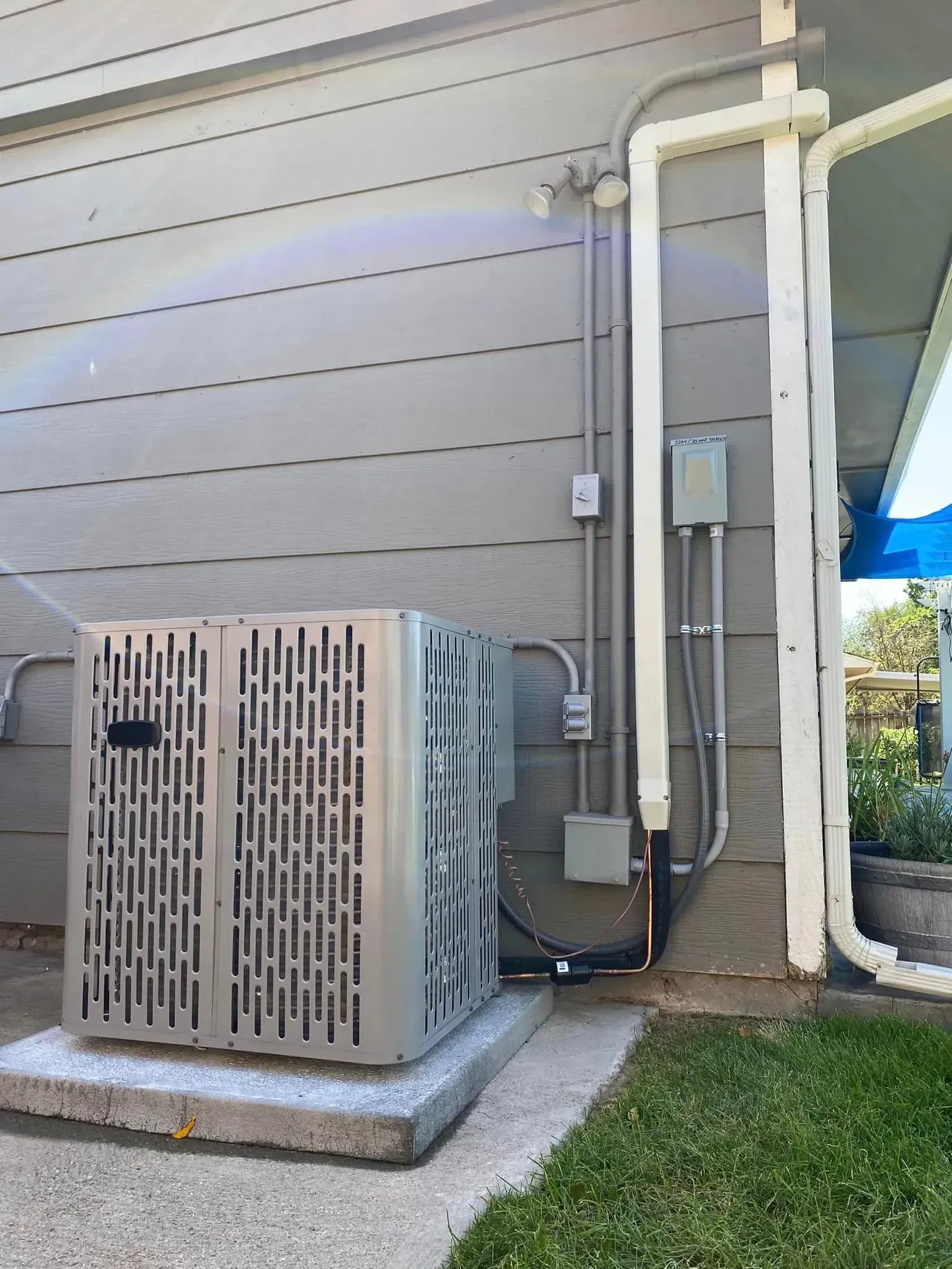 Air conditioner unit next to a gray house with electrical conduit and a downspout.