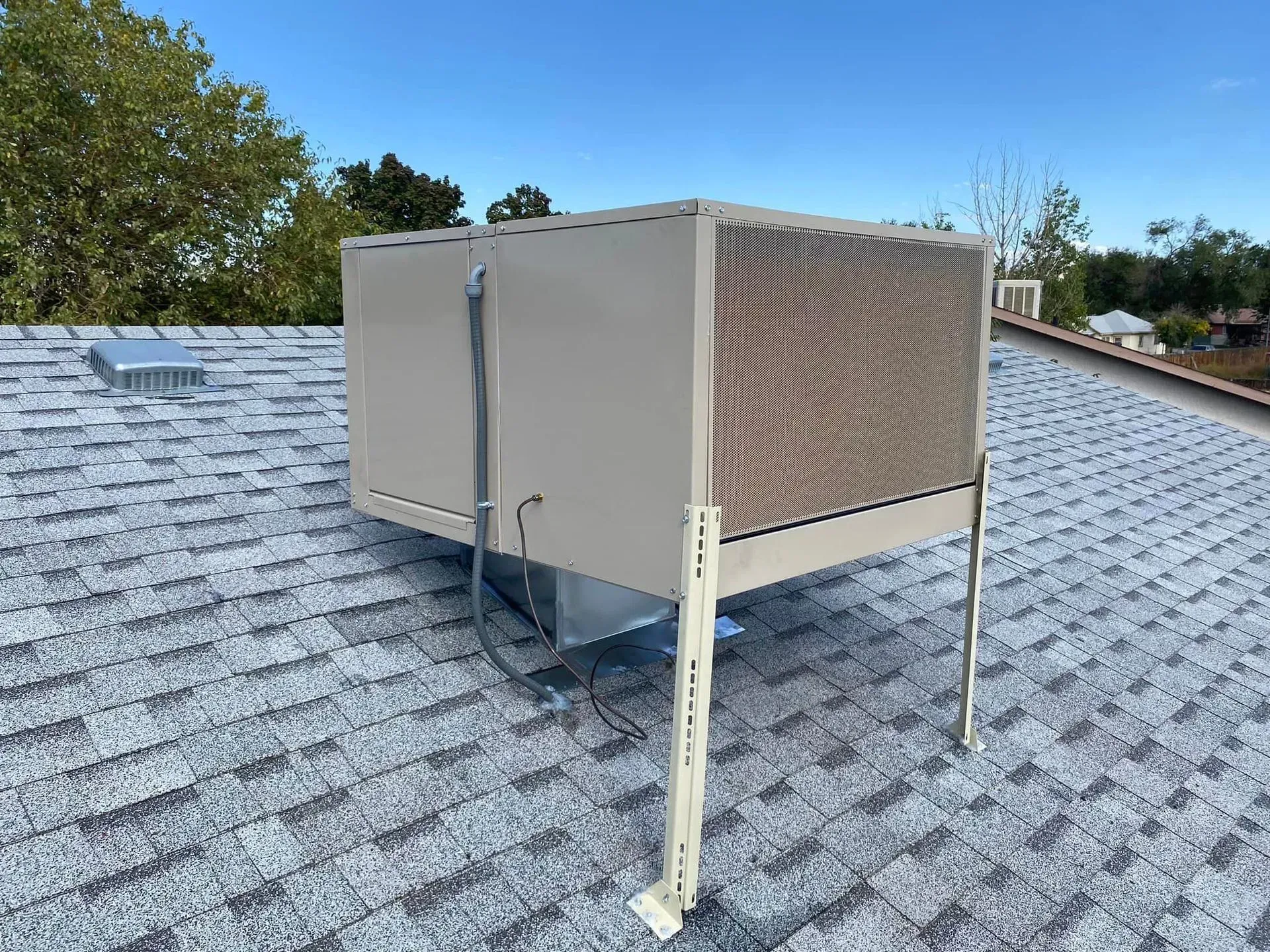 An air conditioning unit on a gray shingled roof against a blue sky.