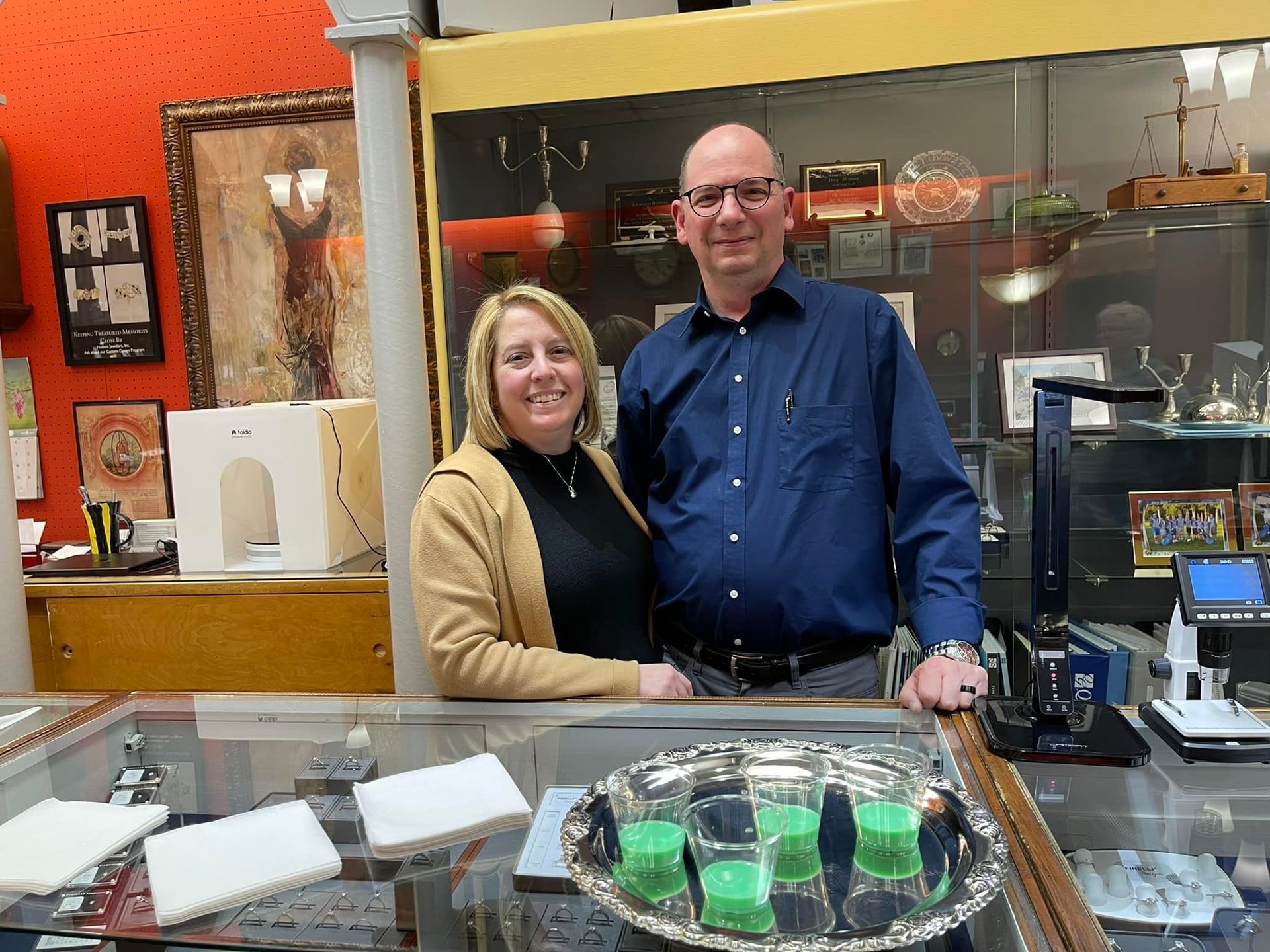 A man and woman stand behind a display case in a store. The man wears a blue shirt; the woman a tan jacket.