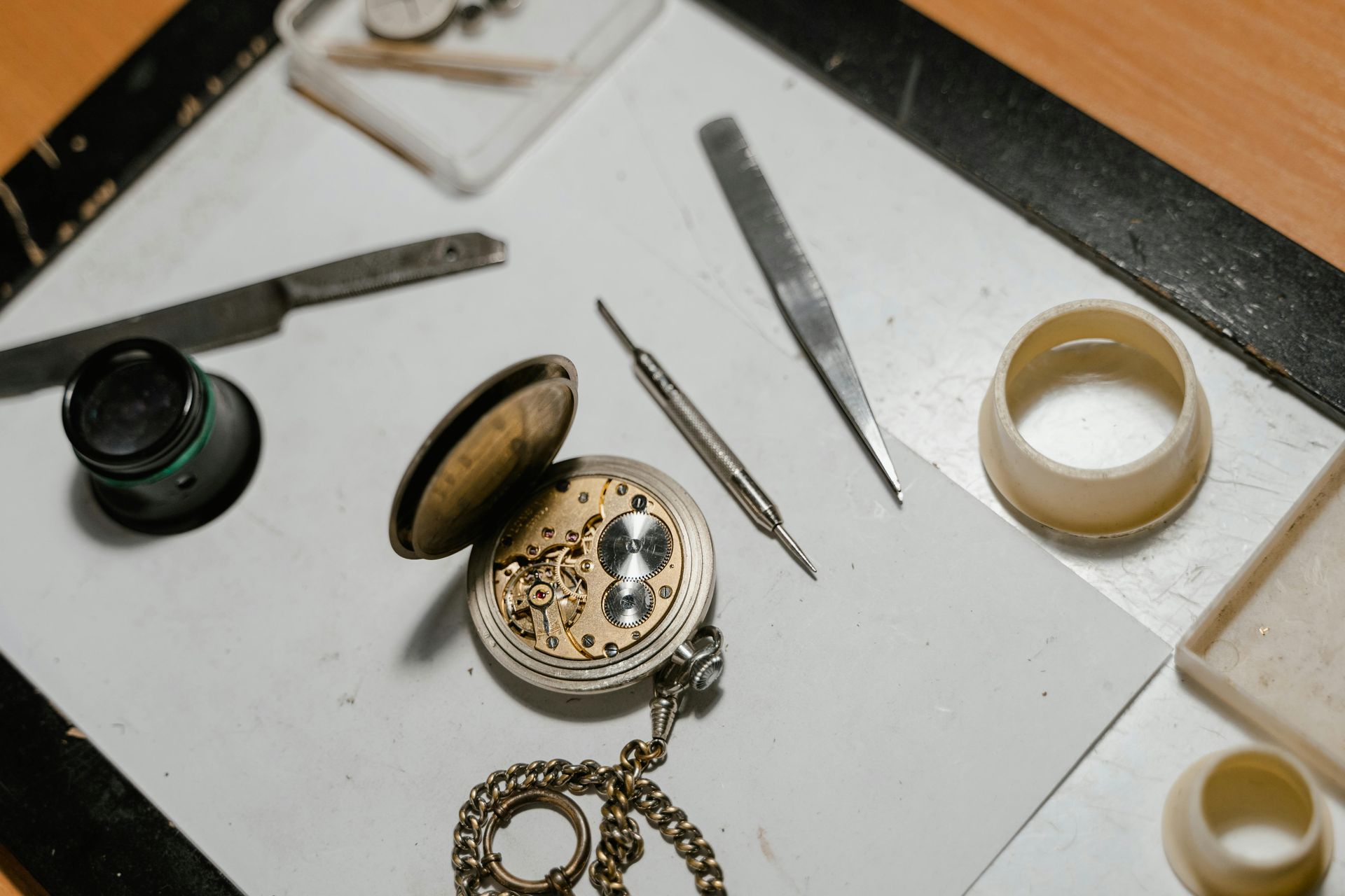 Pocket watch being repaired, with tools like tweezers, knife, and loupe, on a white surface.
