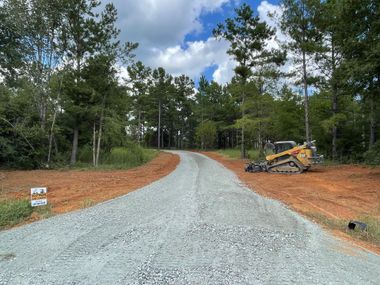 Gravel driveway curves through a wooded area; a bulldozer sits to the side. Blue sky with clouds.