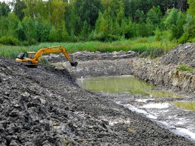 Yellow excavator digging in a muddy pond, surrounded by dirt and trees.