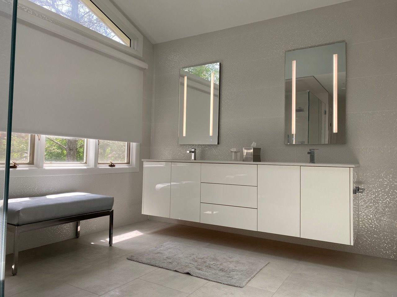 Modern bathroom with floating white vanity, two lighted mirrors, and a window with a shade.