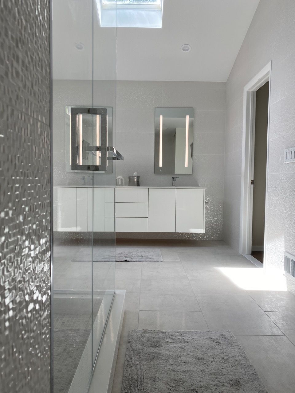 Modern bathroom with floating white vanity, two lighted mirrors, glass shower, and skylight.