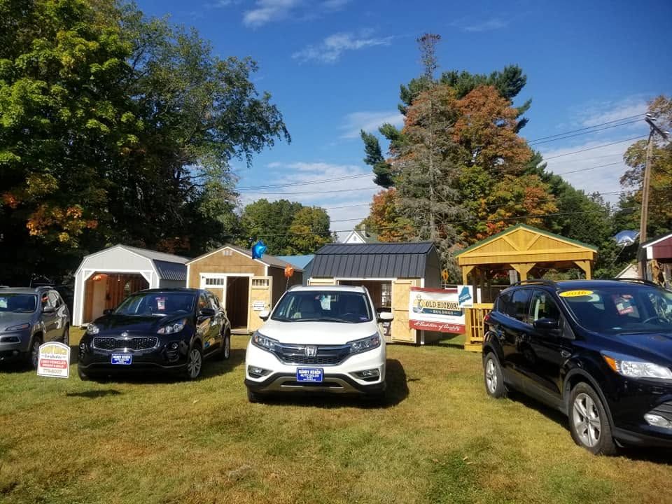 Outdoor car lot with three vehicles parked on grass in front of small cabins under a blue sky | Randy Keach Auto Repair