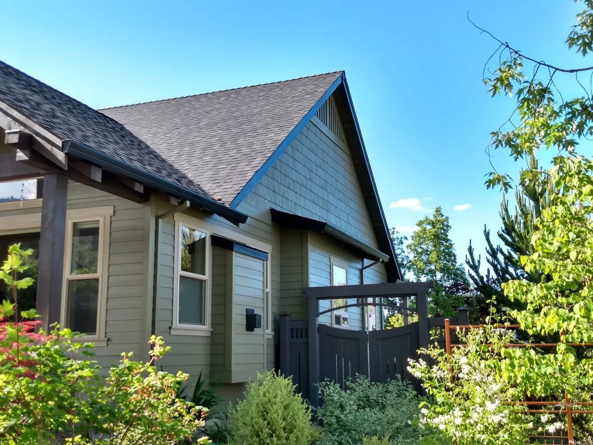 A side view of a green house with dark roof shingles, a wooden gate, and lush green landscaping against a blue sky.