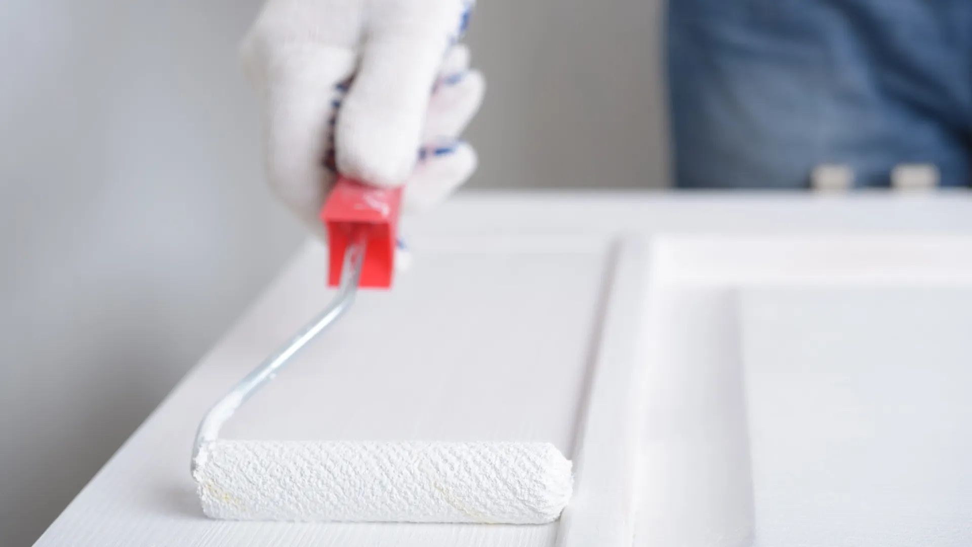 A hand in a white glove uses a small paint roller to apply white paint to a wooden cabinet door.