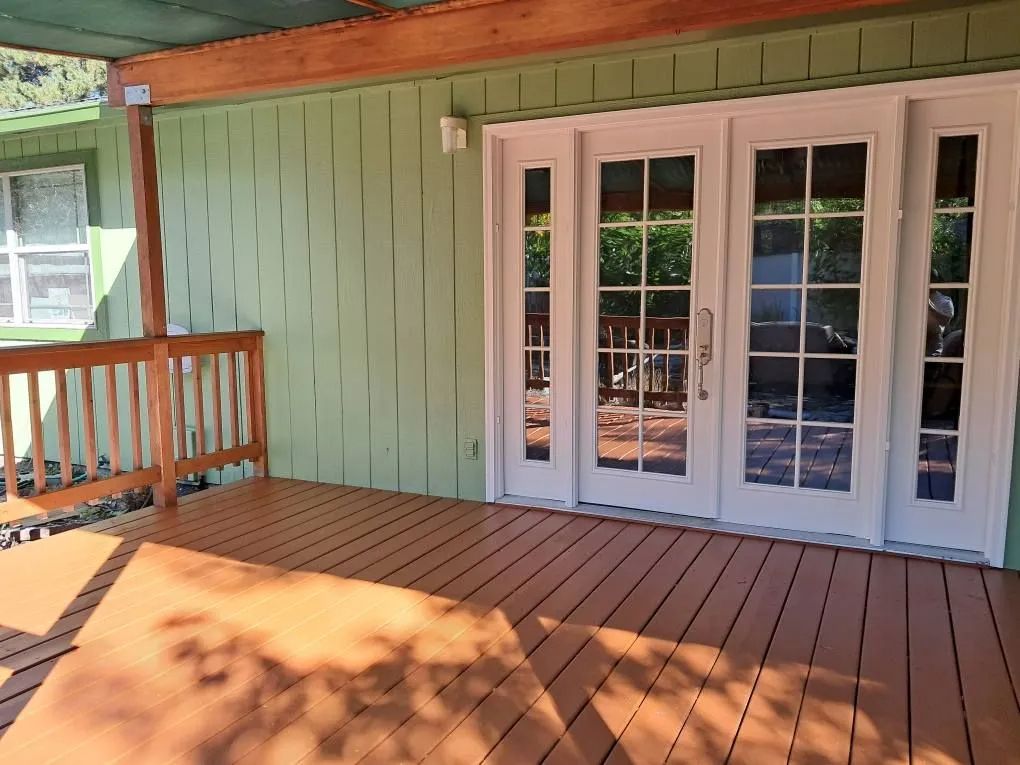 A light green house exterior with a wooden deck, a wooden railing, and a white French door with side panels.