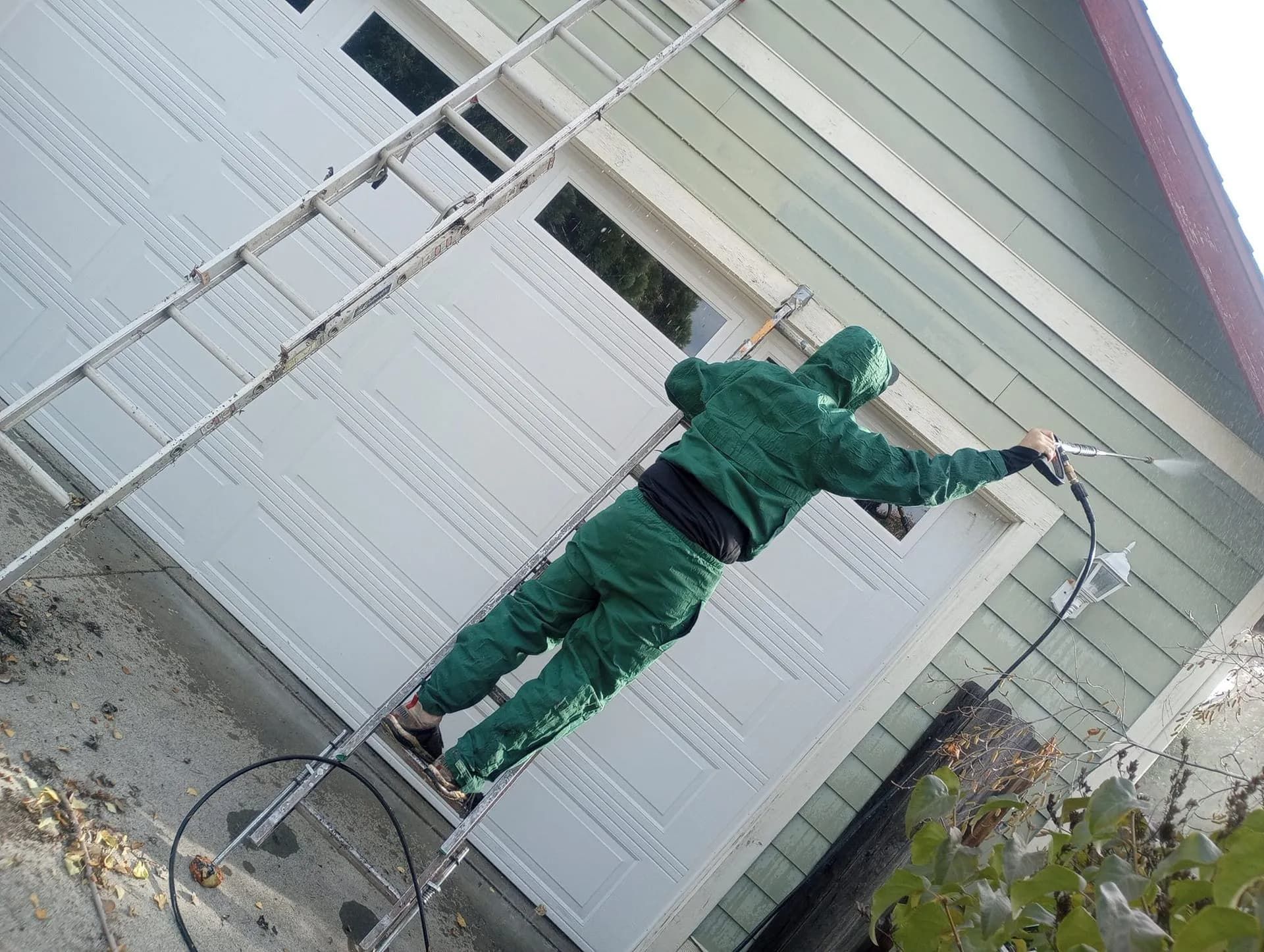 A person in a green jumpsuit on a ladder uses a sprayer to paint a house exterior white.