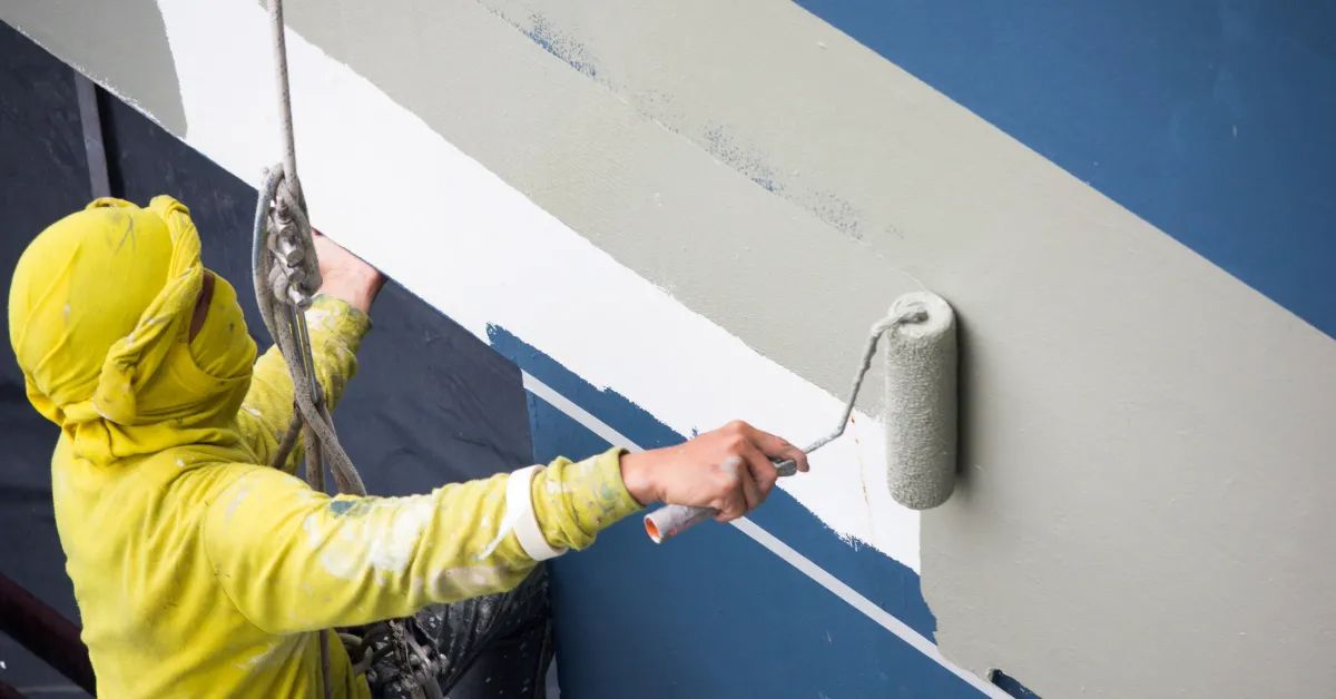 A person in a yellow uniform uses a paint roller to apply light gray paint to a blue exterior wall.