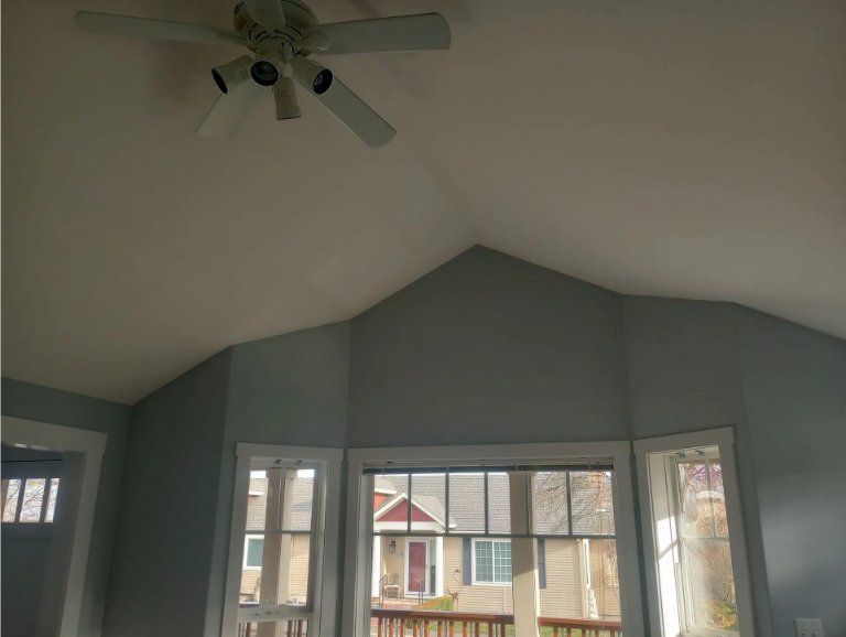 A vaulted ceiling room with a white ceiling fan and blue walls, featuring a bay window overlooking a house exterior.