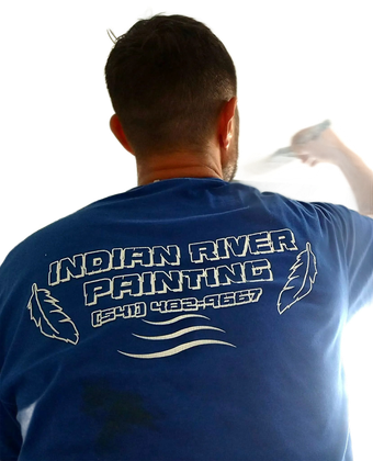 A person wearing a blue hard hat and work overalls, reaching up to use a paint roller against an invisible wall.