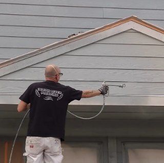 A person wearing a black shirt sprays light-colored exterior paint onto a house with an airless sprayer.