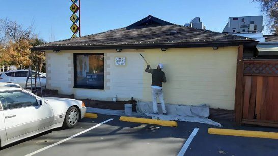 A painter in light clothing works on the exterior wall of a building, painting the light-colored brick with a roller.