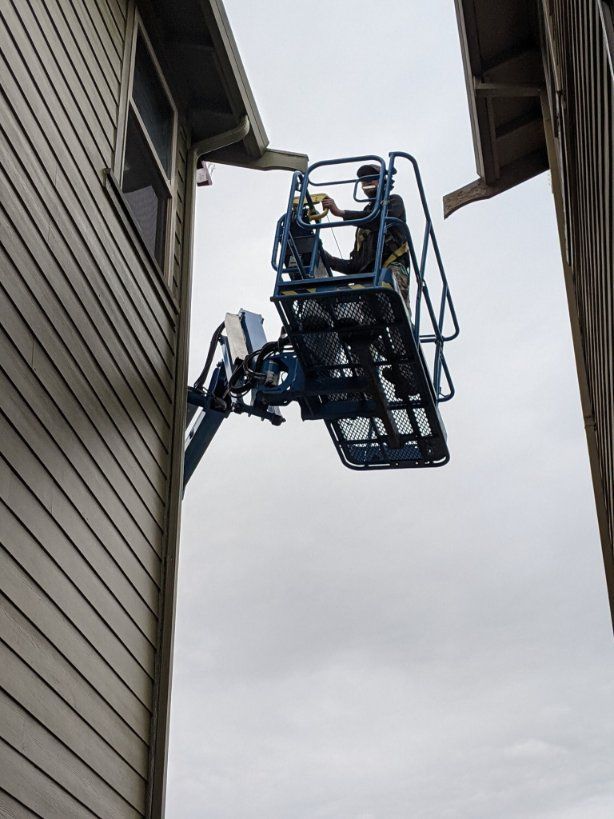 A worker in a blue boom lift reaches toward the roof gutter of a beige siding house.