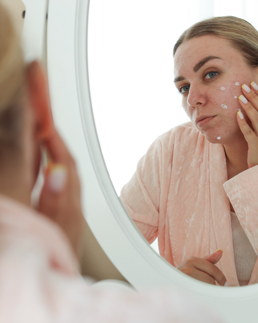 Person applying cream to face in mirror, wearing a light pink robe