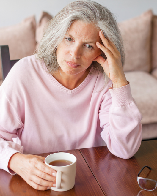 Woman in a pink sweater sitting at a table, holding a mug and resting her head thoughtfully.