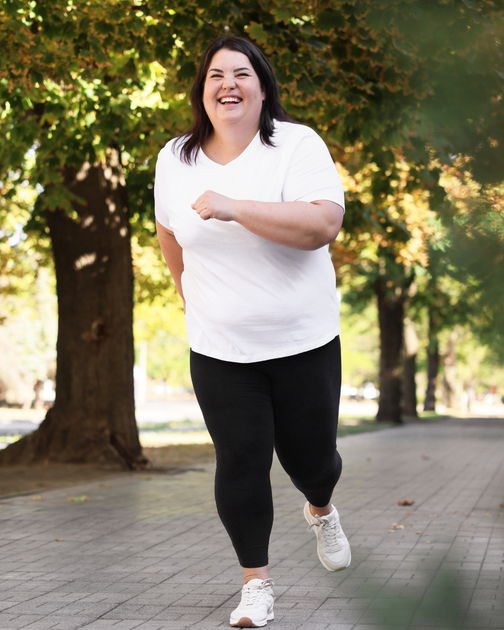 Woman jogging on a tree-lined path in a white shirt and black leggings, smiling outdoors.