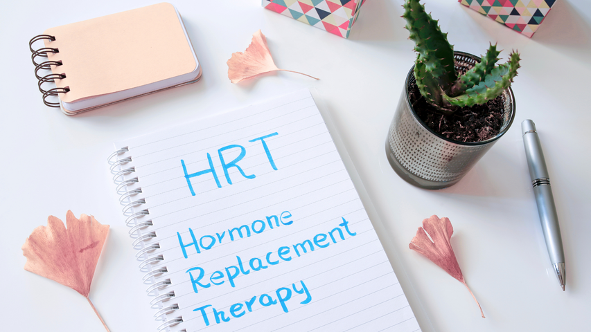 Notebook with “HRT Hormone Replacement Therapy” on a desk, beside a plant, pen, and pastel stationery.