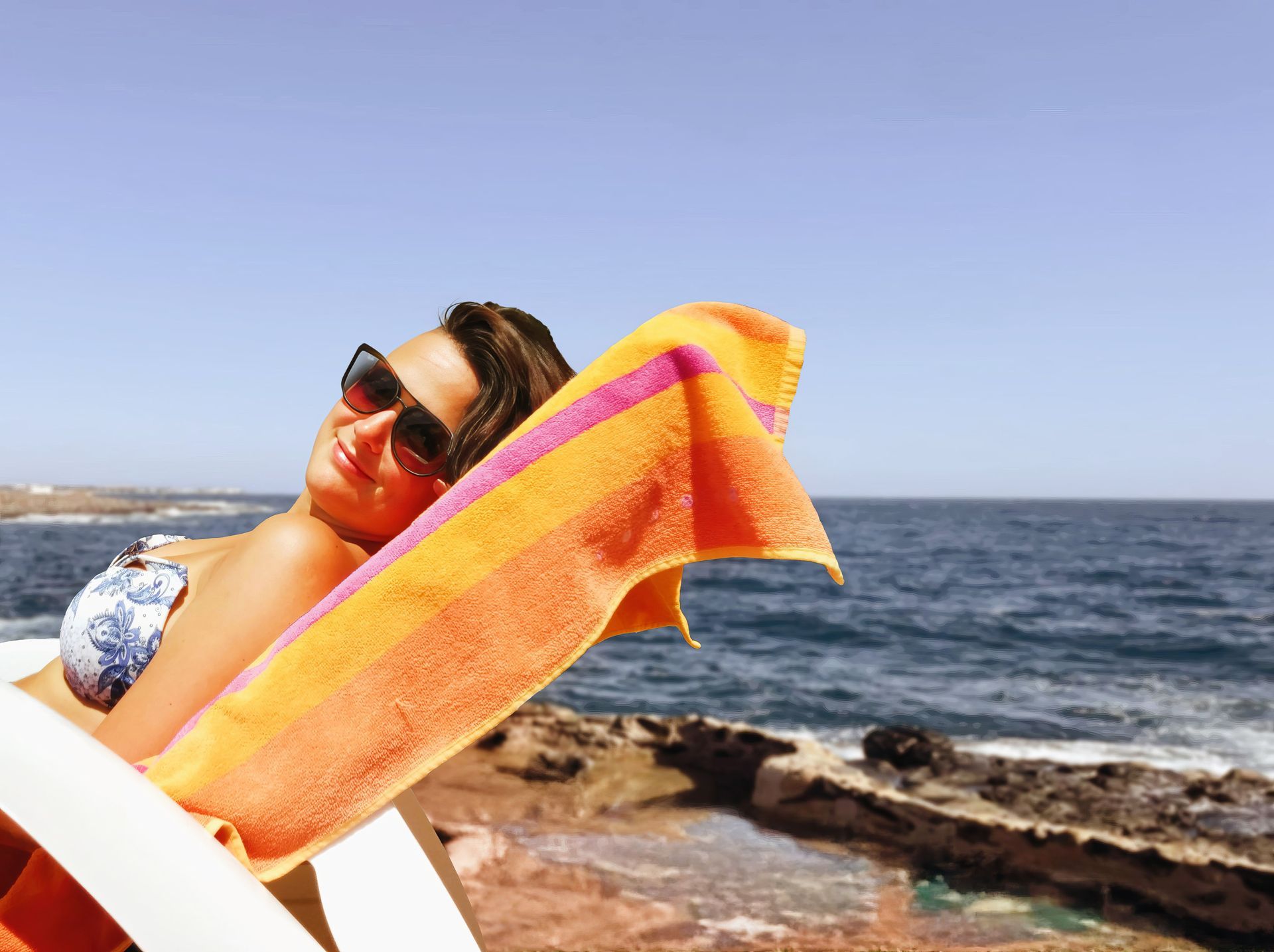 Frau mit Sonnenbrille liegt entspannt auf einer Sonnenliege an der Felsküste am Meer