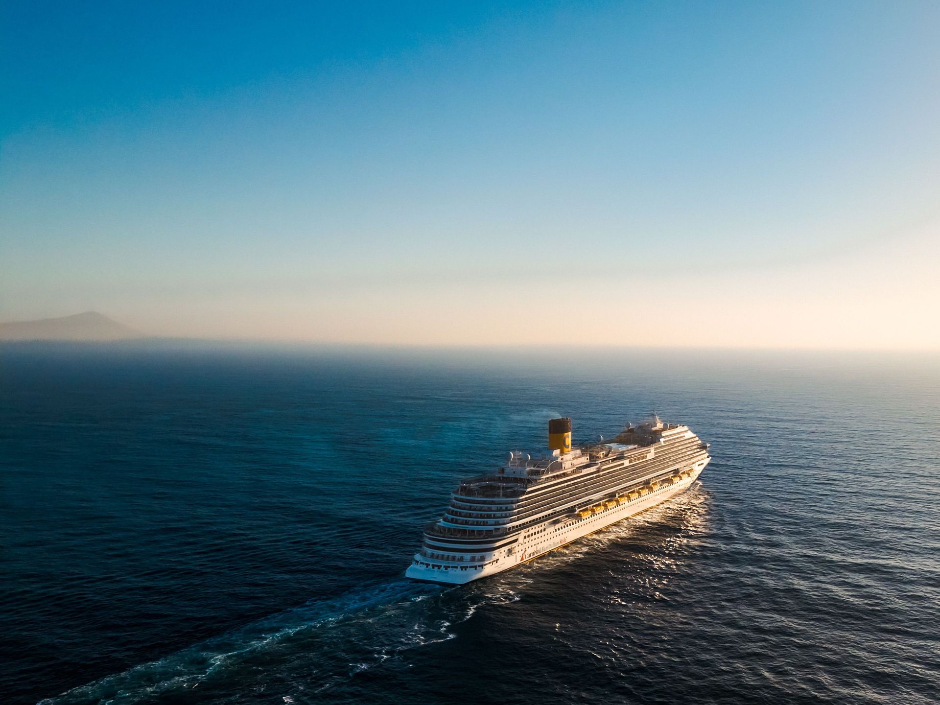 Großes Kreuzfahrtschiff auf offener See, von der Sonne beschienen, mit langer Kielspur im Wasser.