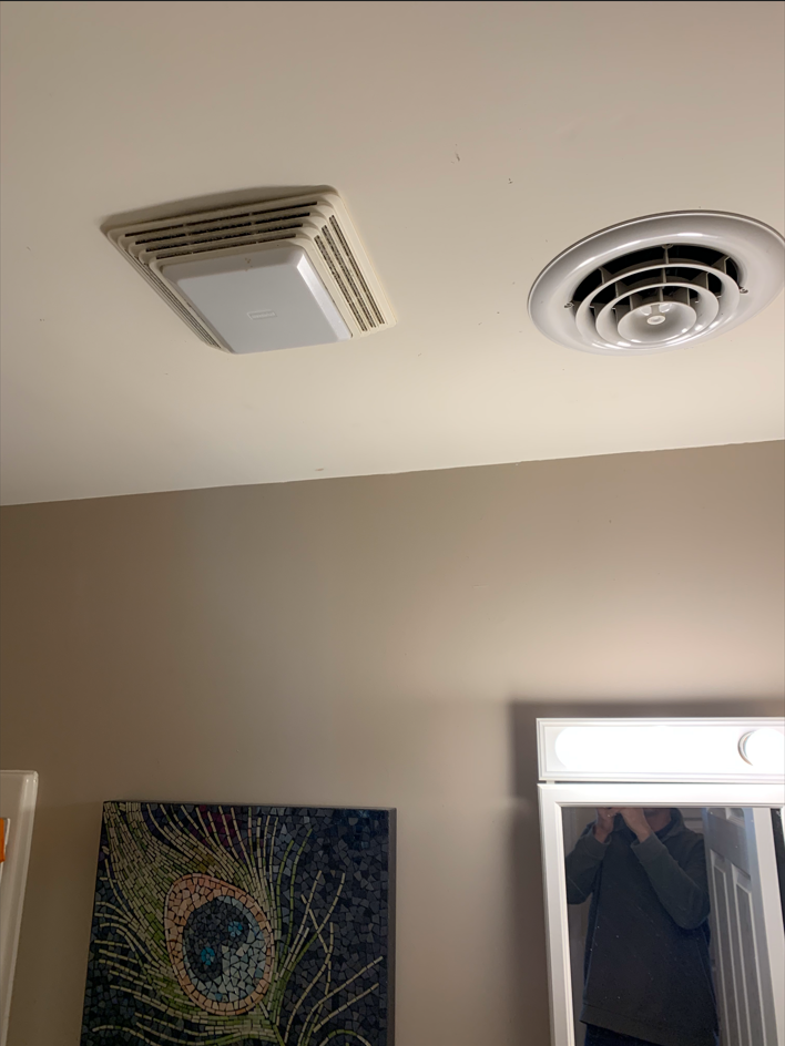 Ceiling with a vent and round air vent above a beige wall and lit mirror