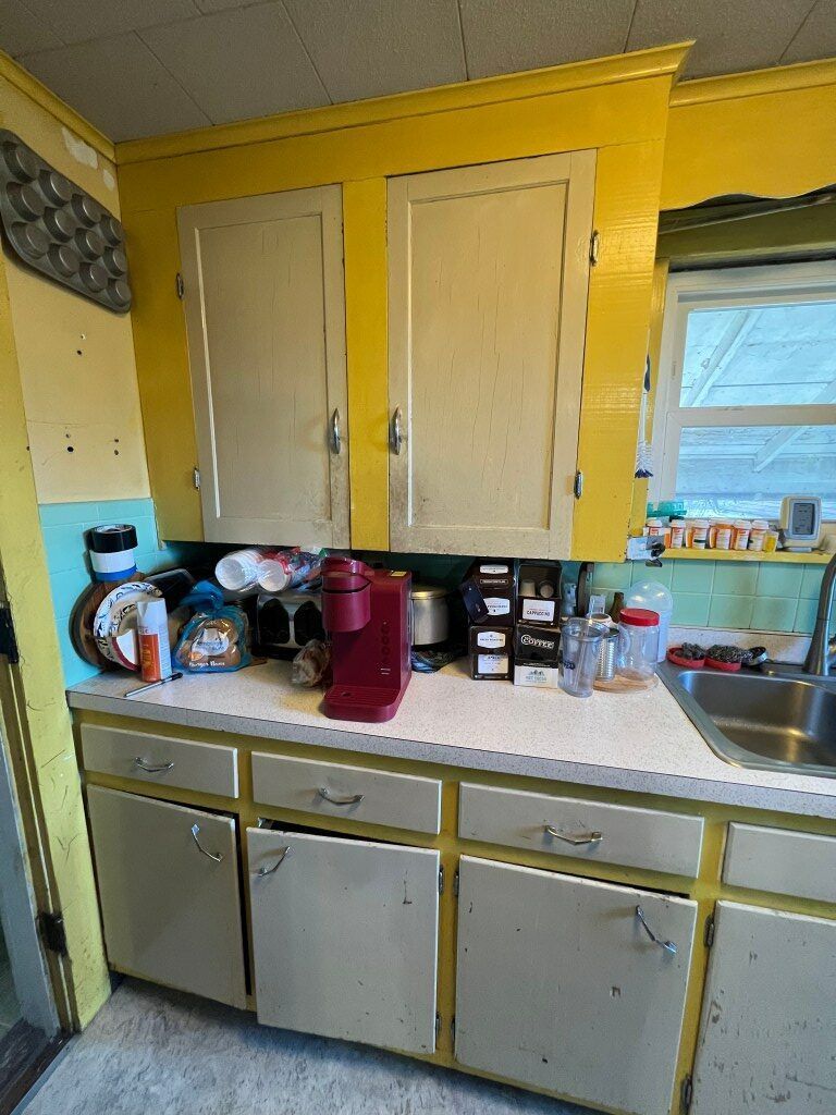 Yellow kitchen with white cabinets, cluttered countertop, sink, and window above the counter.