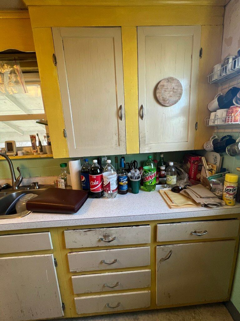 Yellow kitchen cabinets and cluttered countertop with assorted bottles and dishes under a cabinet.
