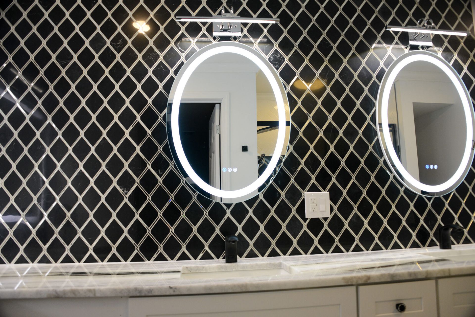 Two illuminated oval mirrors above a bathroom counter with black-and-white diamond tile wall.