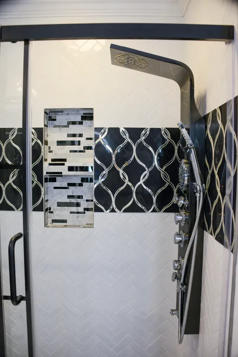Glass shower door with a black curved grab bar and black-and-white mosaic tile strip on the wall