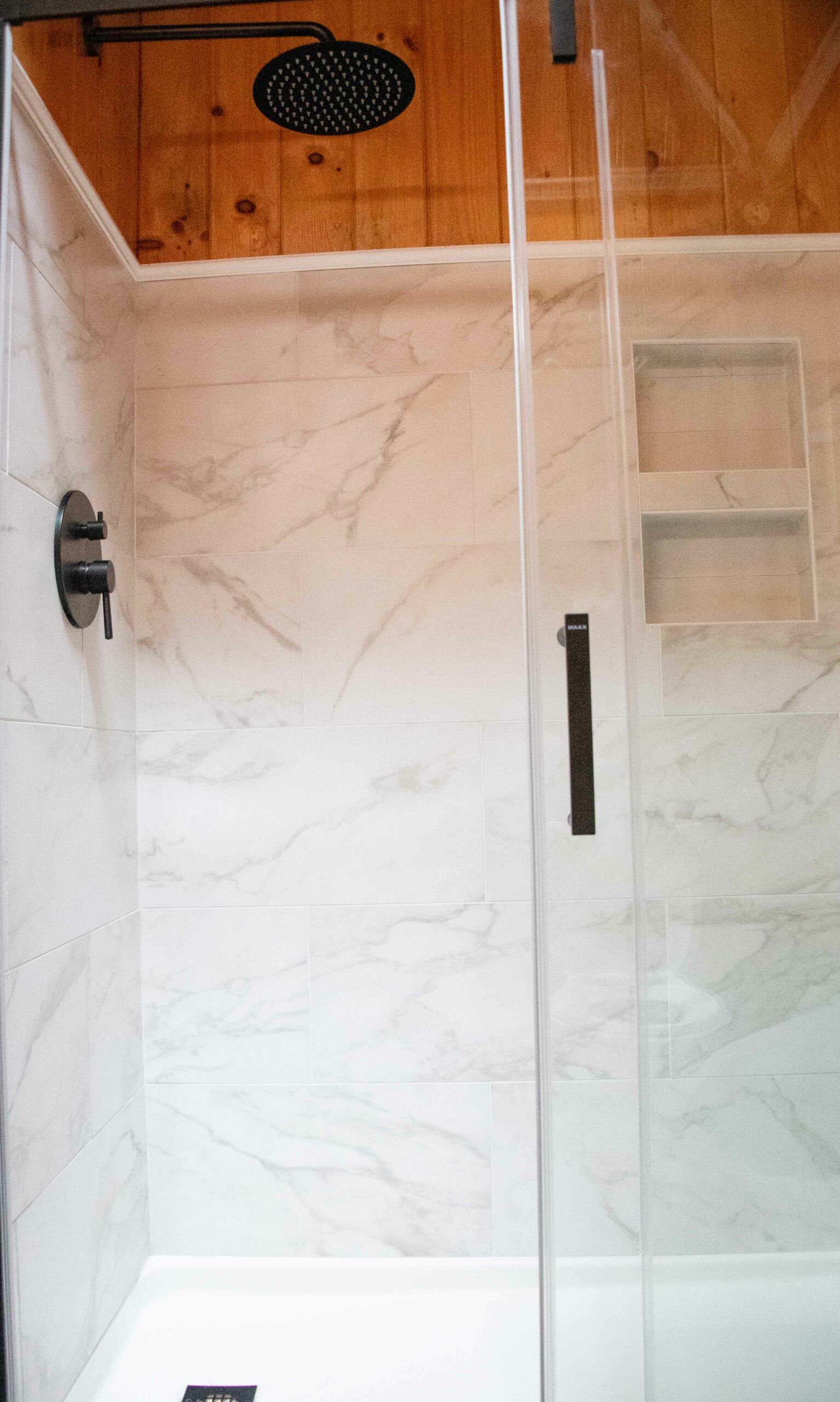 Walk-in shower with beige tile, glass door, black showerhead, and built-in corner shelves.