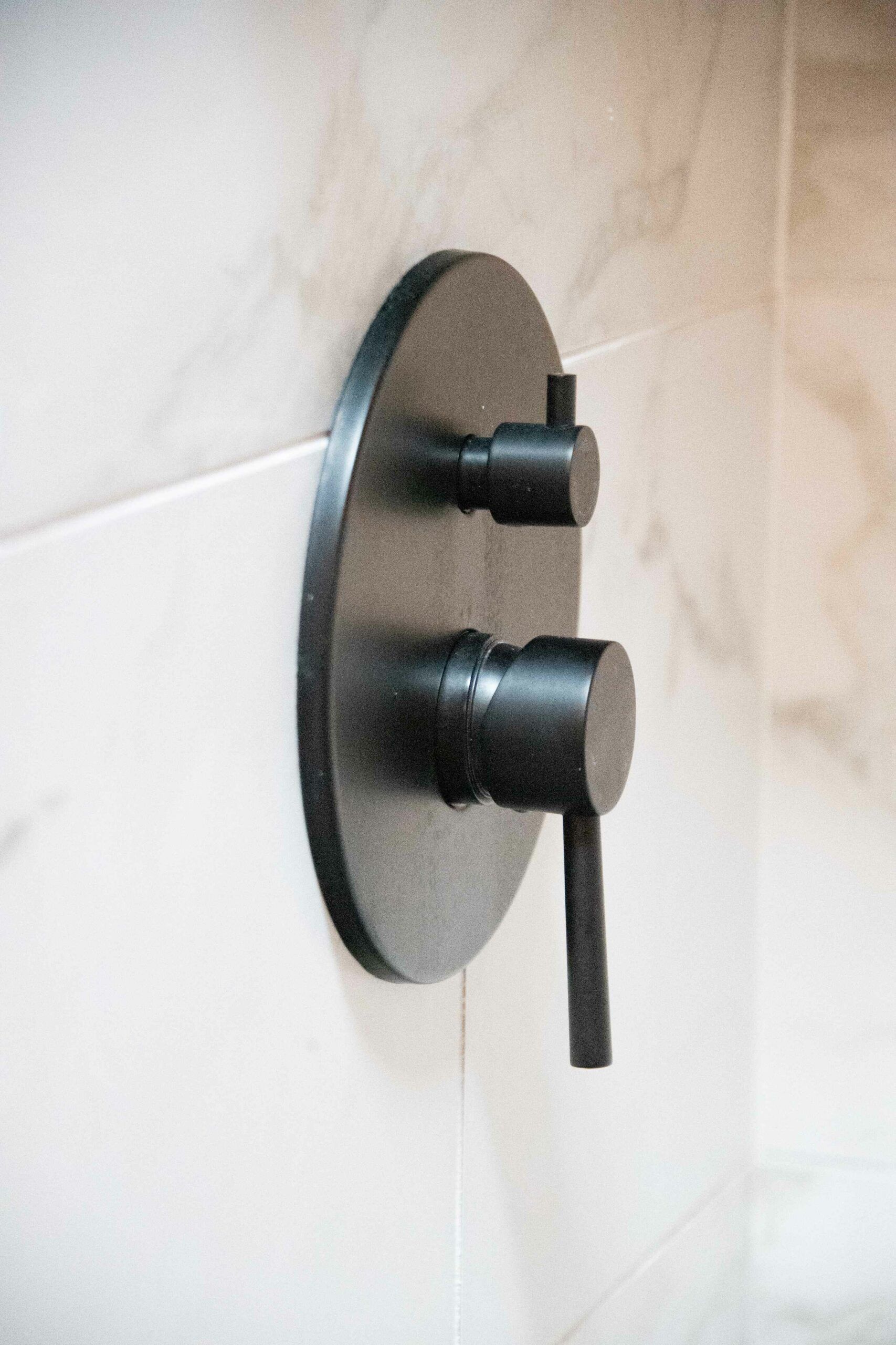 Black shower control panel with two knobs on a light tiled wall