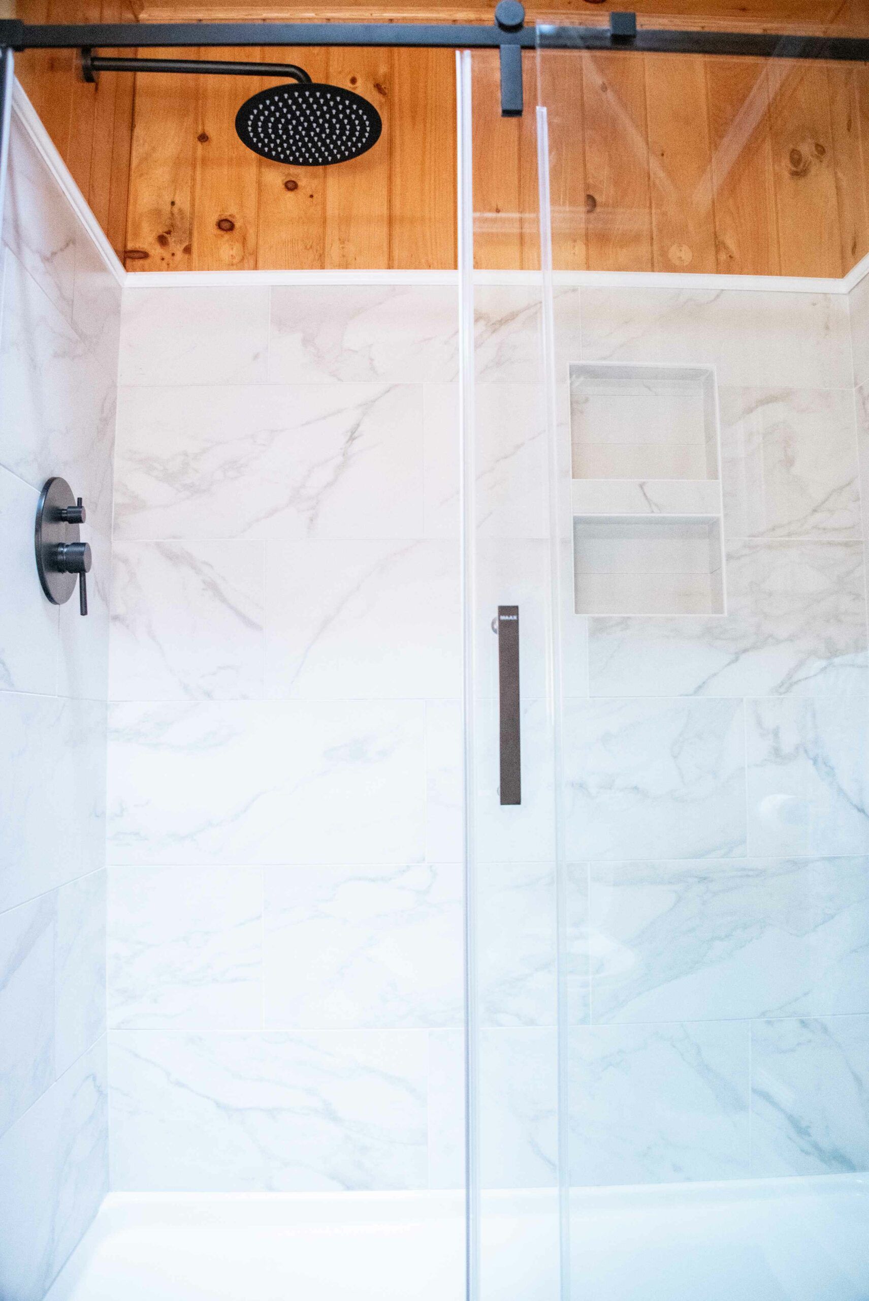 Glass shower with marble tile walls and a black rainfall showerhead underneath a wood ceiling.
