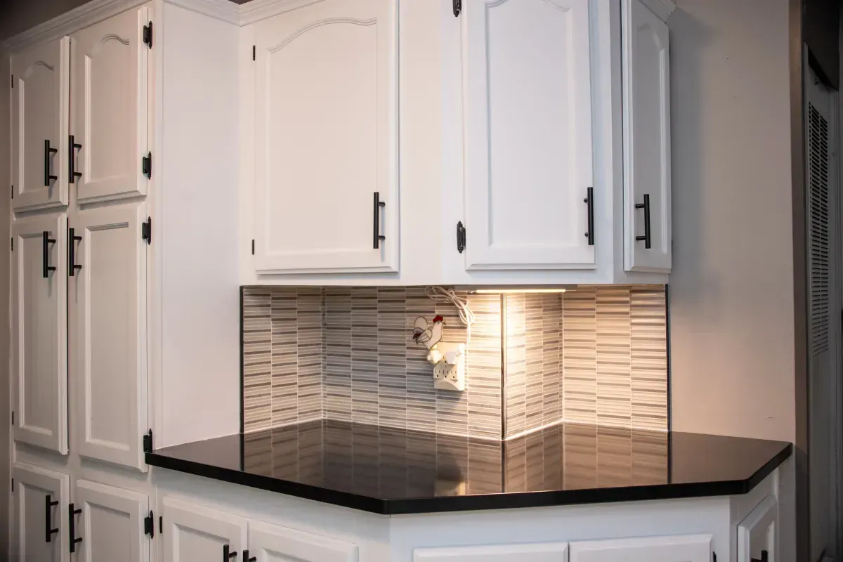 White kitchen cabinets with dark countertops and a tiled backsplash corner under a cabinet light