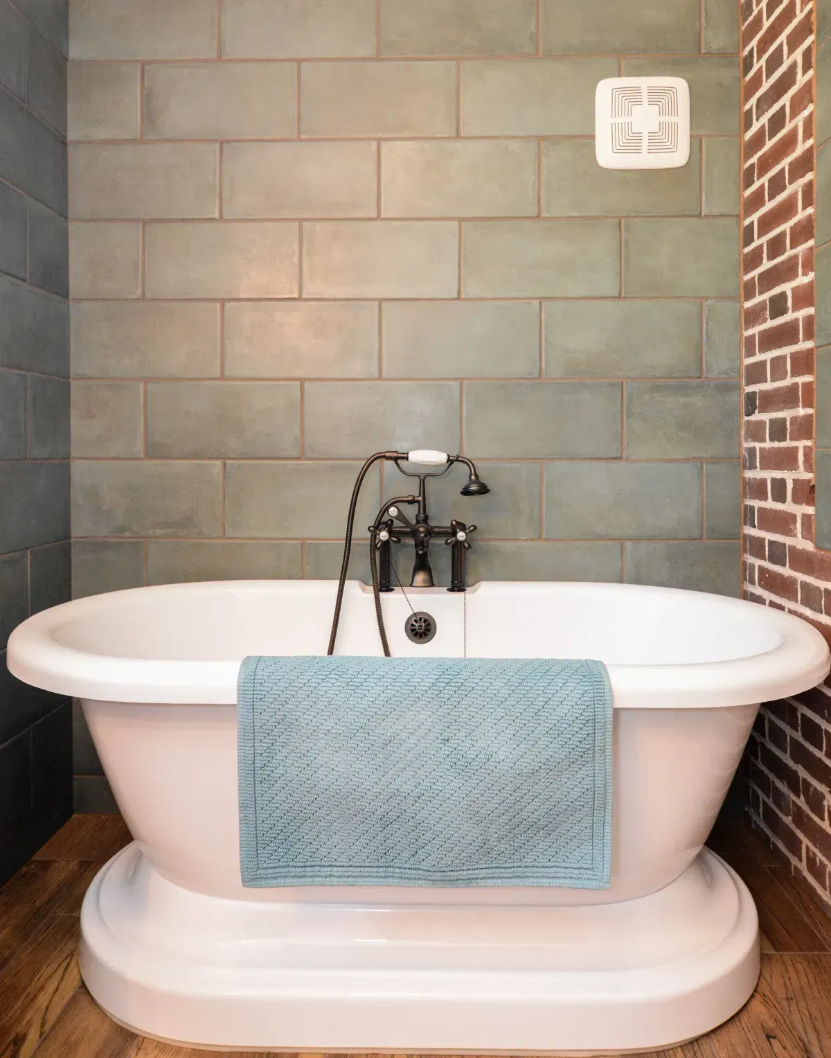 White bathtub with faucet and blue towel against gray tile and brick bathroom walls