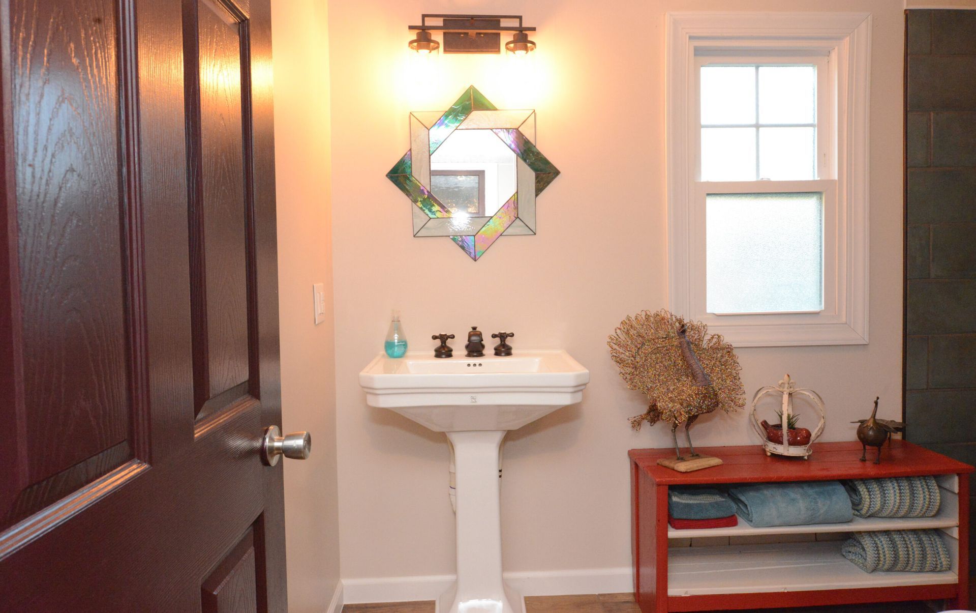 Small bathroom with pedestal sink, wall mirror, window, and a red storage bench.