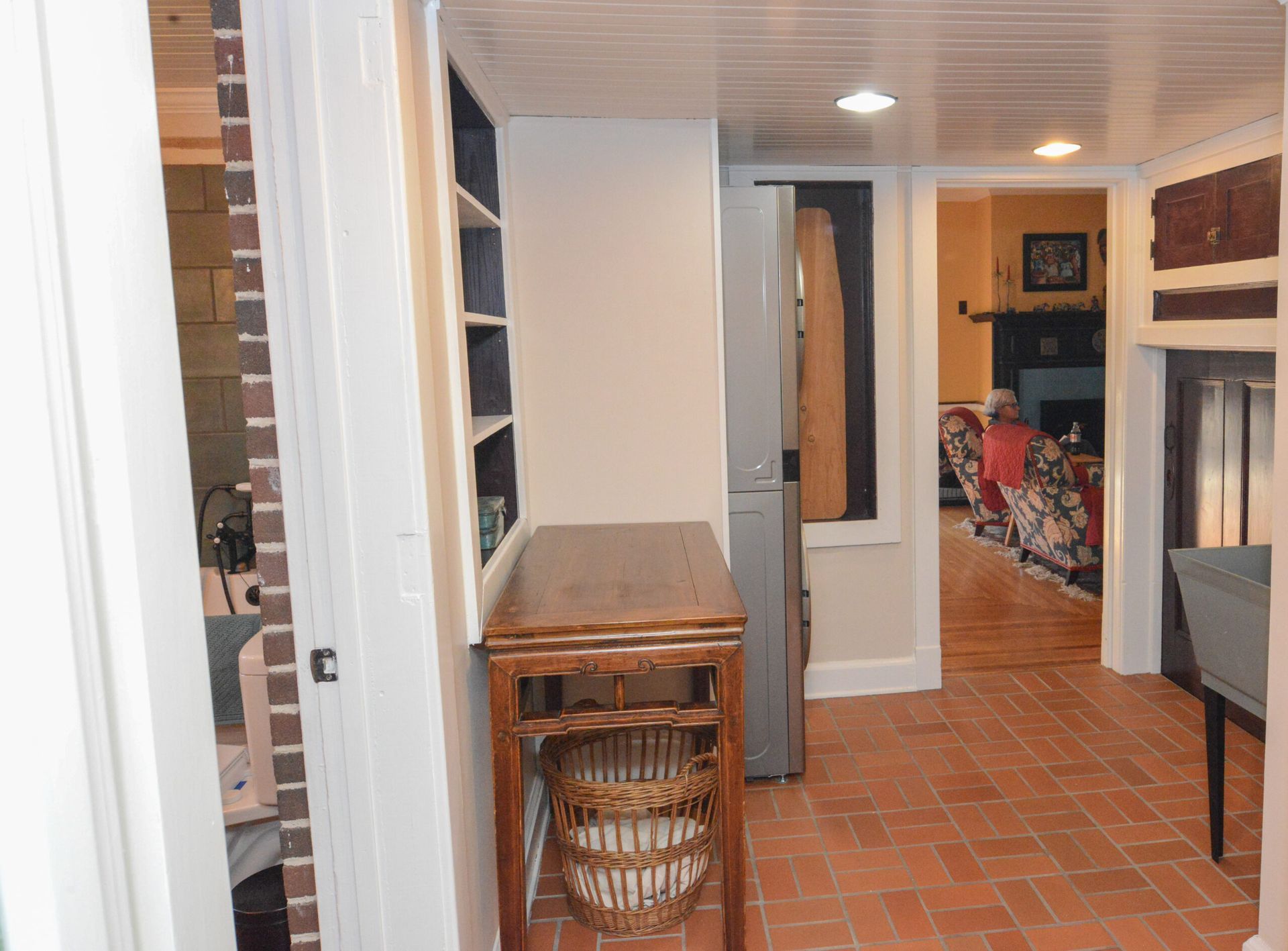 Narrow hallway with terracotta tile floor, wicker table, shelving, and an open view into a dining area.