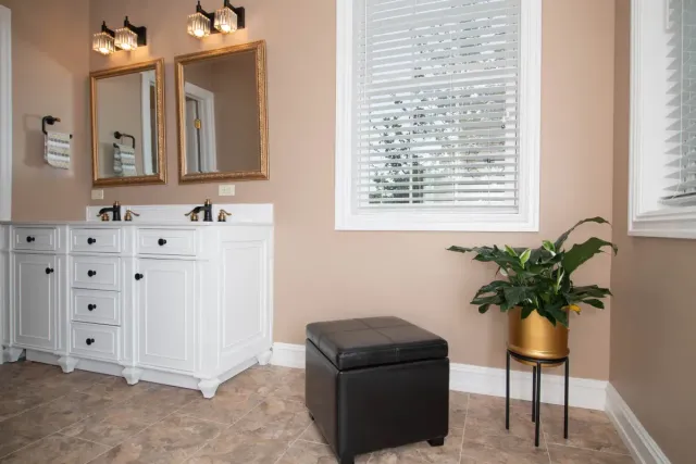 Bright bathroom with white vanity, twin mirrors, window blinds, black ottoman, and potted plant.