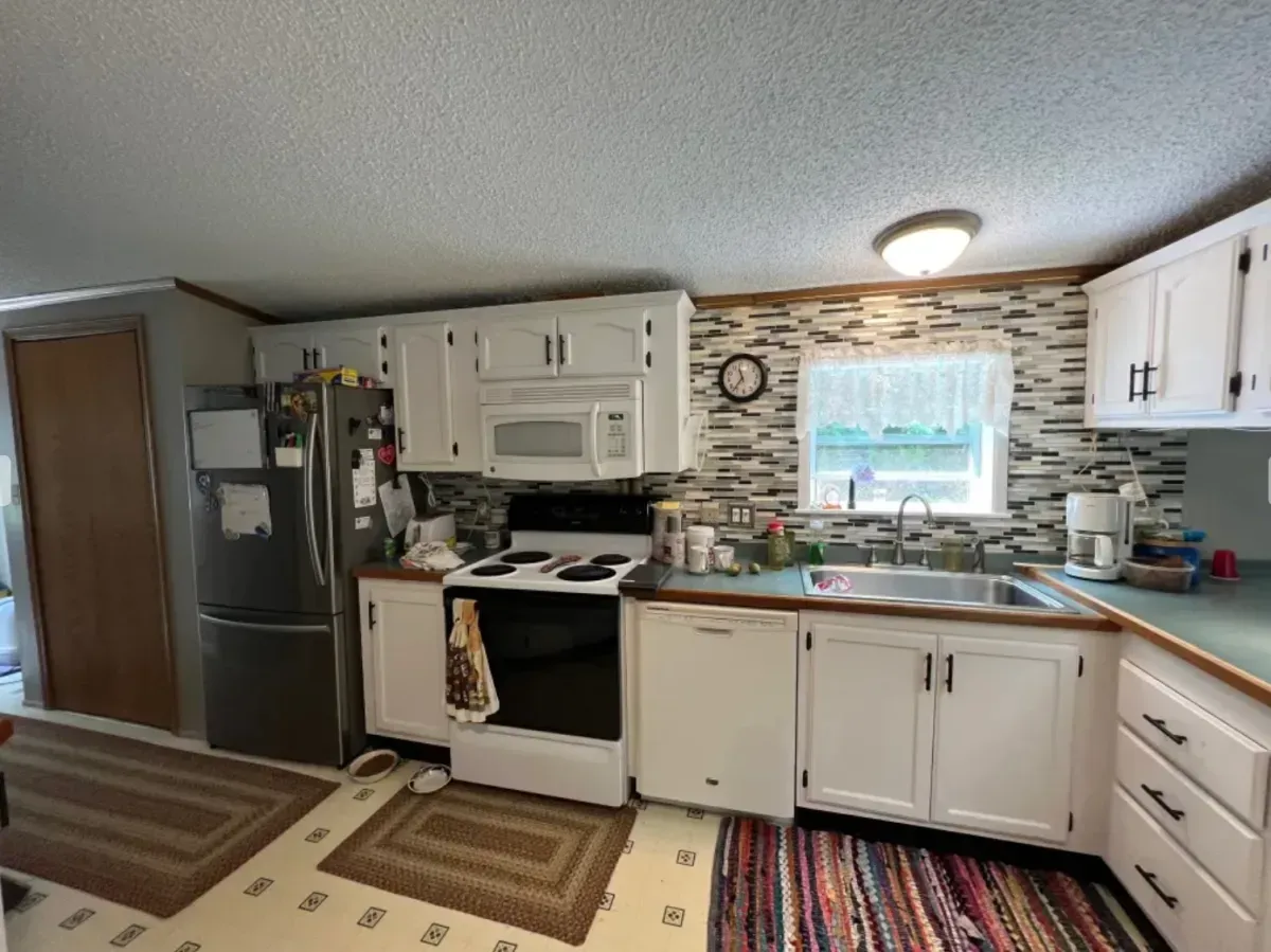 Kitchen interior with white cabinets, stainless fridge, stove, sink, and mosaic backsplash under a window