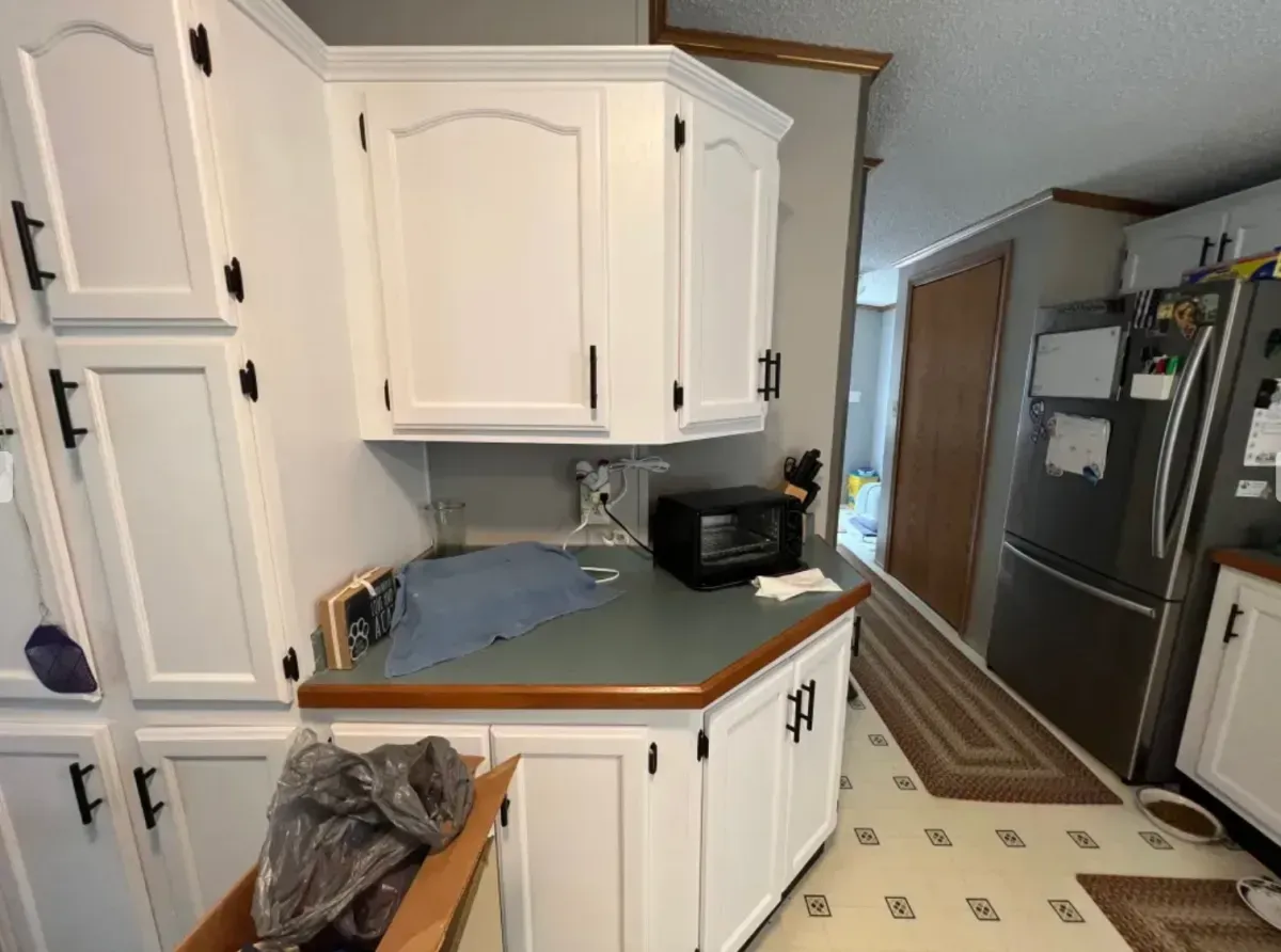 White kitchen with corner cabinets, black countertop, stainless fridge, and a hall rug leading to a doorway.