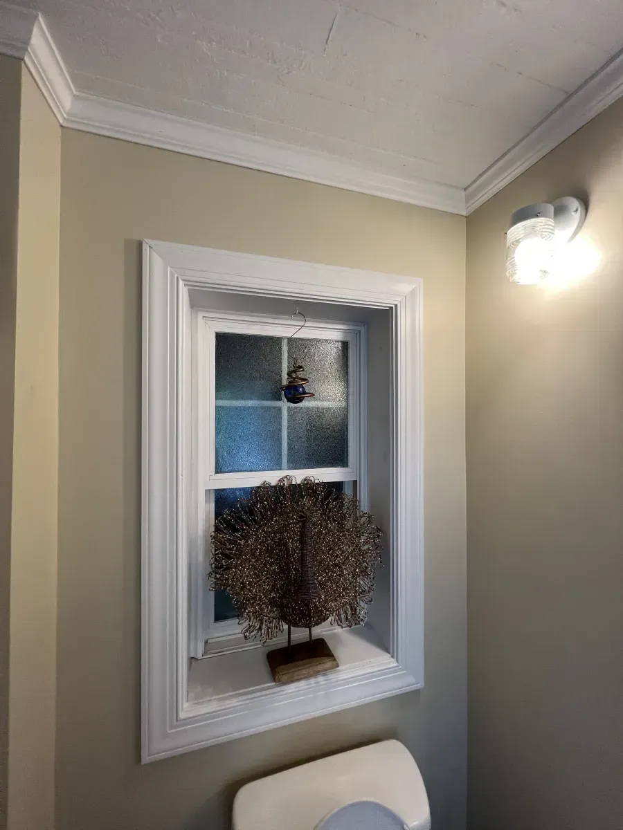 Framed window with a potted plant on the sill in a beige hallway, lit by a wall sconce.