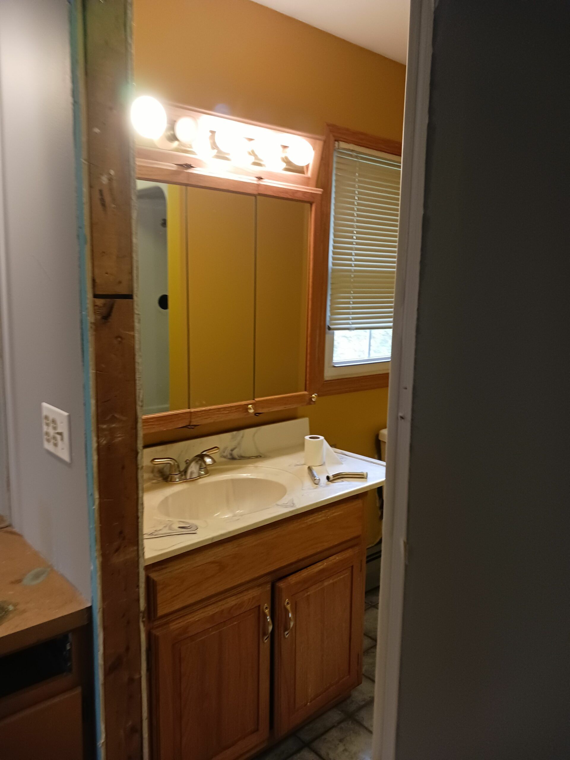 Bathroom vanity with wood cabinets, mirror, and lit light fixture beside a window with blinds