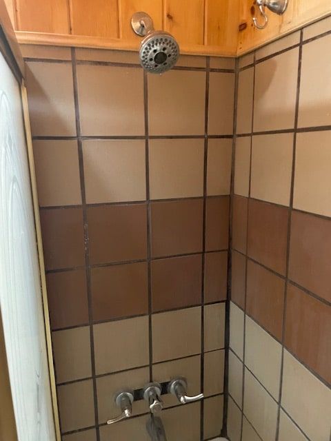 Small tiled shower corner with brown and beige tiles, chrome showerhead, and wooden cabinet above.