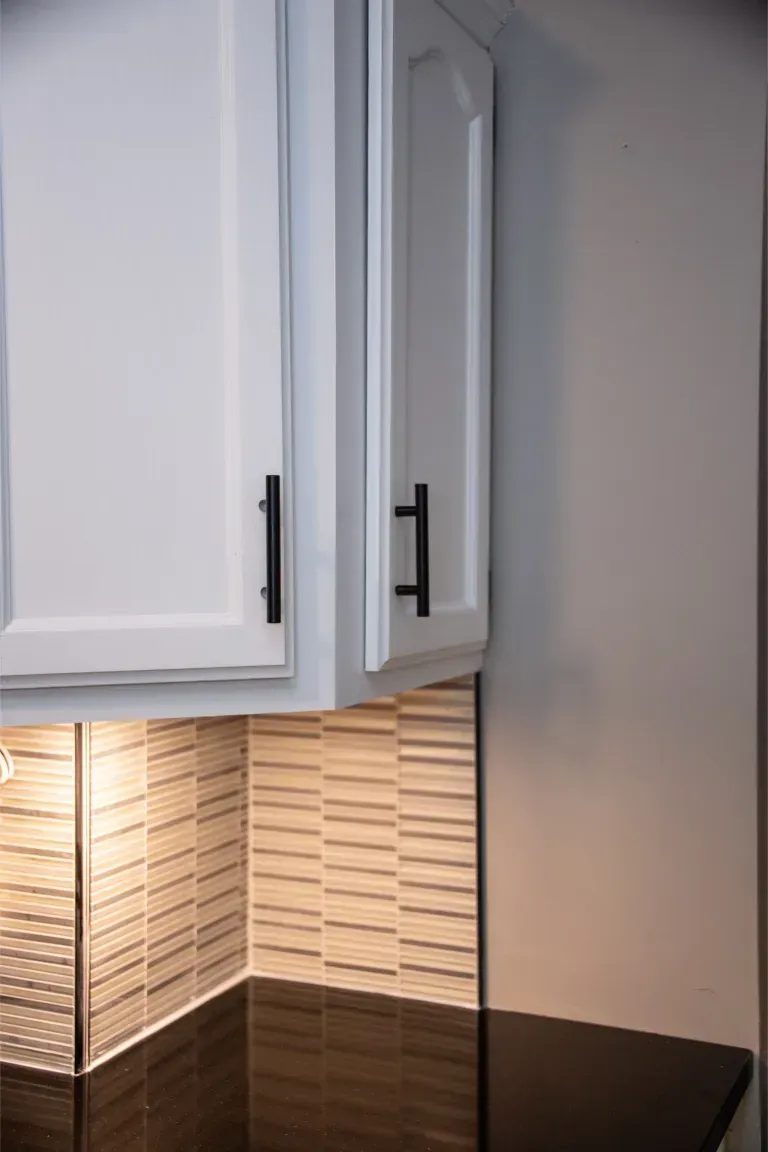 Kitchen cabinets with white doors and black handles above a lit tile backsplash.