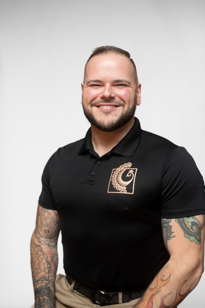 Smiling man in a black polo shirt with tattooed arms standing against a light gray background
