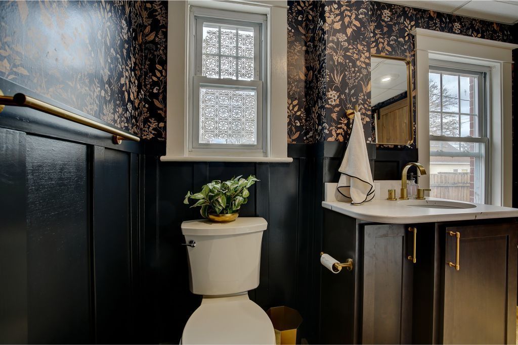 Small bathroom with black wainscoting, patterned wallpaper, toilet, sink, and mirror by a window.