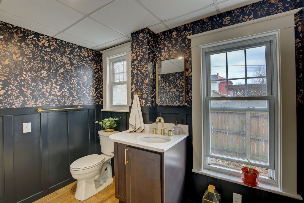 Bathroom with dark paneling, floral wallpaper, toilet, vanity sink, and two windows with a waterfront view
